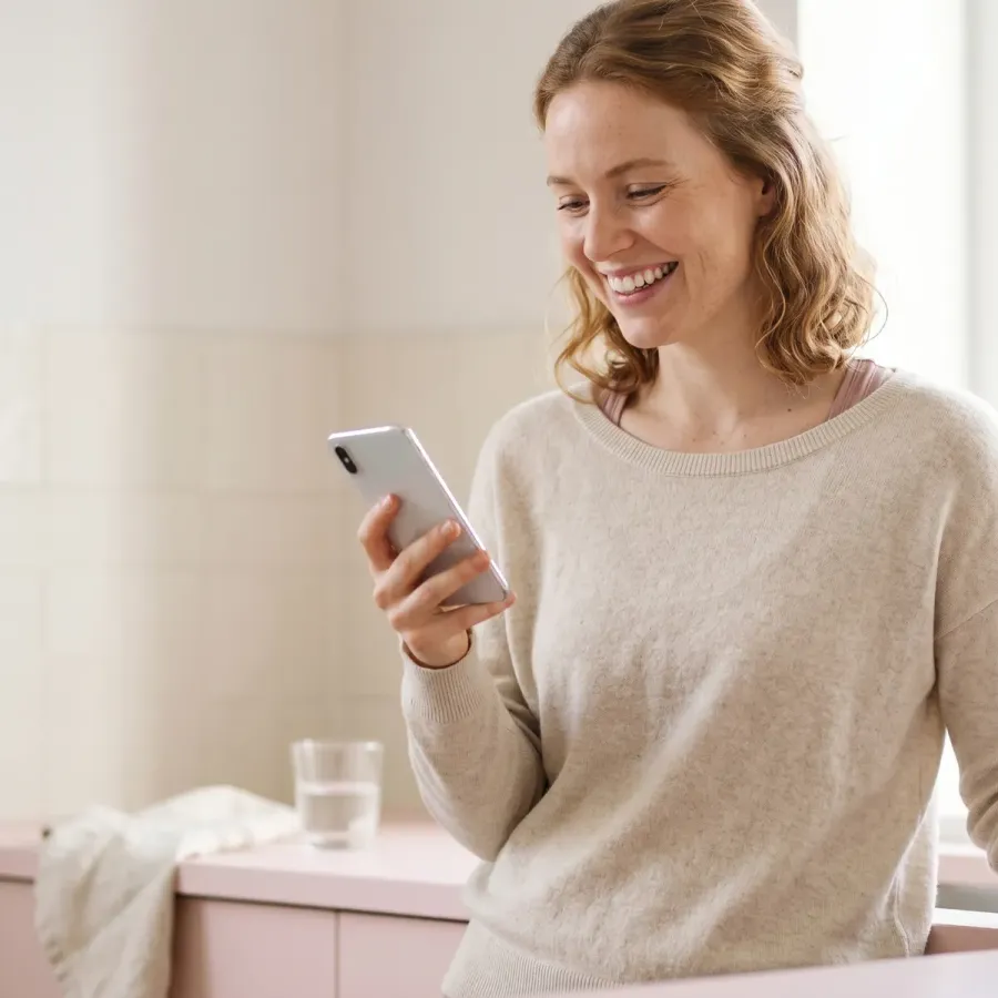 Woman in a cream sweater smiling at her phone in a soft pink kitchen during a standard telehealth consult